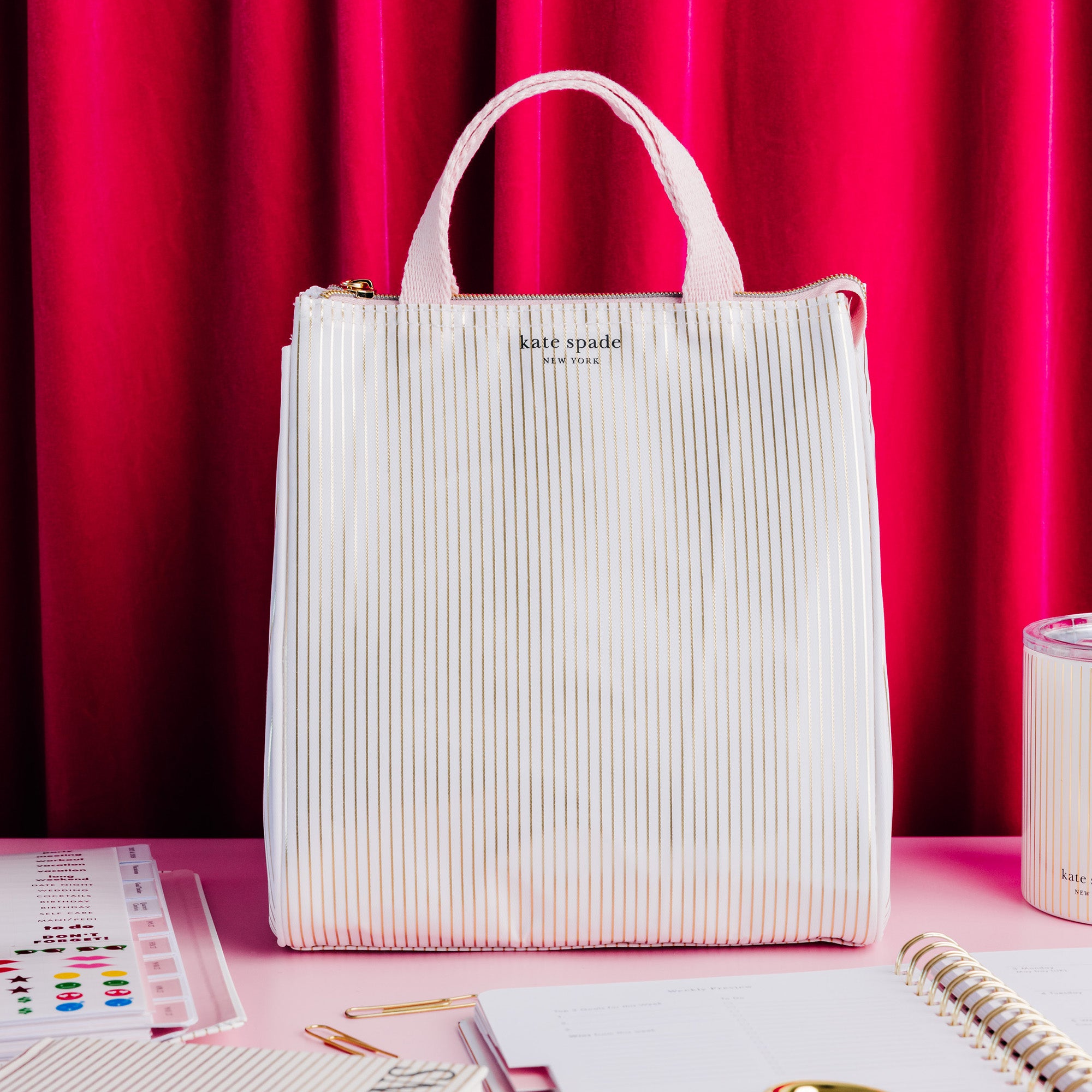 A Kate Spade New York lunch  bag in a textured, vertically pleated off-white fabric, set against a vibrant pink and red curtain backdrop with stationery props on a pink table.