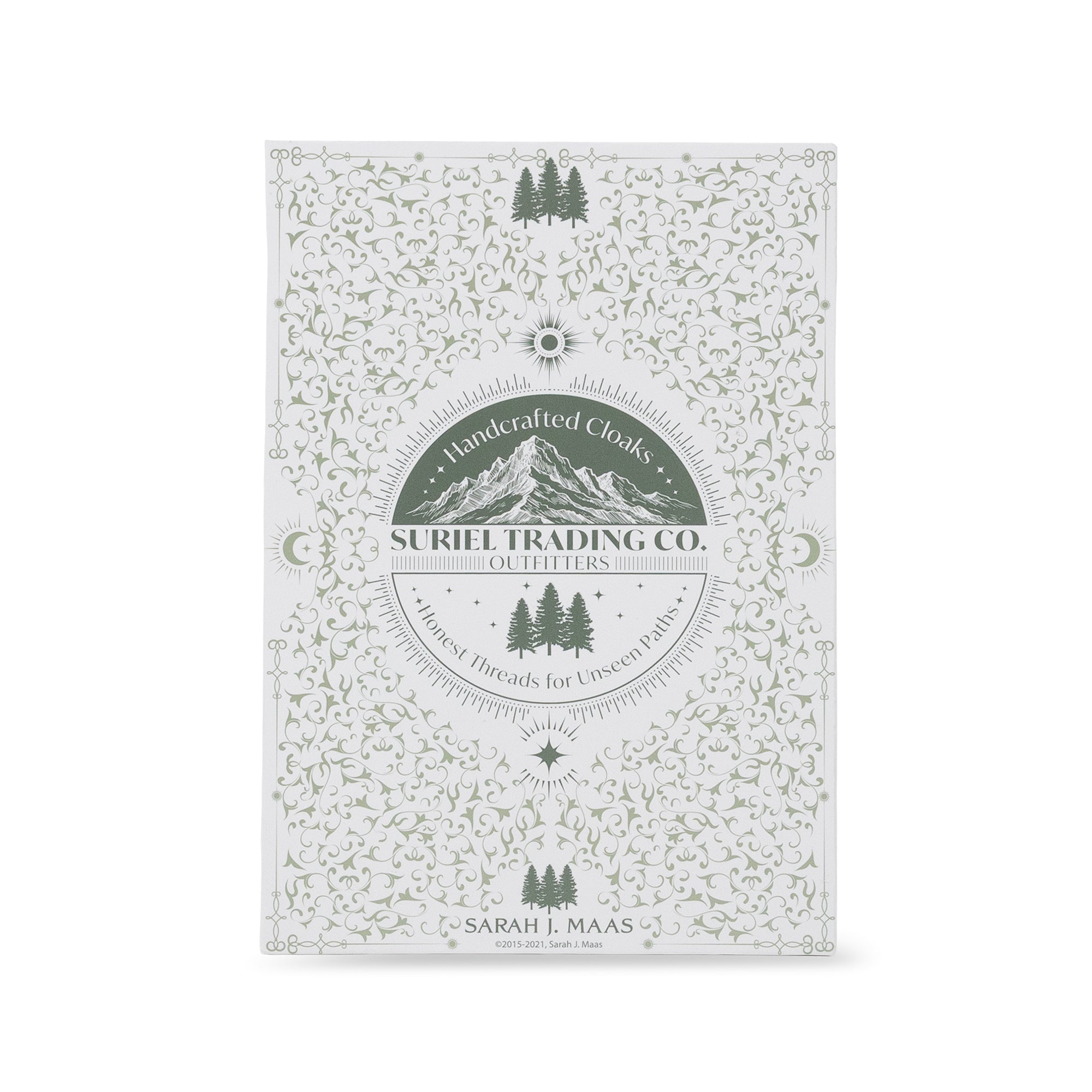 suriel trading co. embossed gold ornament with gold hanging ribbon book packaging with ornate light green floral and vine patterns, featuring a central circular label that reads 'Handcrafted Cloaks SURIEL TRADING CO. OUTFITTERS' with mountain and tree graphics, photographed on a clean white background.
