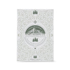 suriel trading co. embossed gold ornament with gold hanging ribbon book packaging with ornate light green floral and vine patterns, featuring a central circular label that reads 'Handcrafted Cloaks SURIEL TRADING CO. OUTFITTERS' with mountain and tree graphics, photographed on a clean white background.