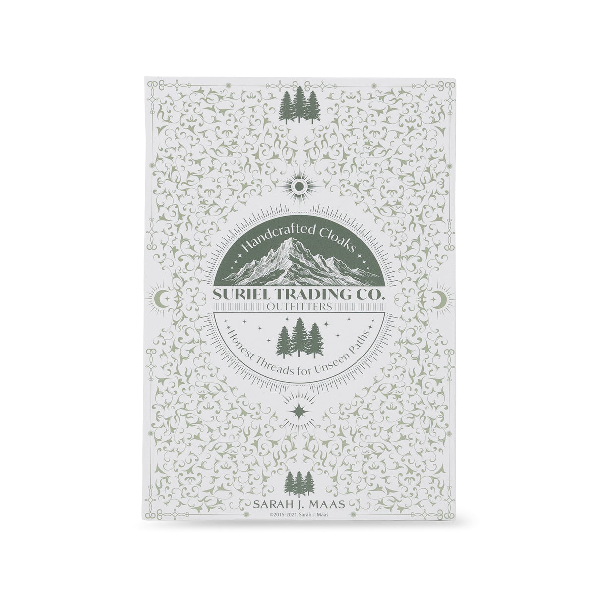 suriel trading co. embossed gold ornament with gold hanging ribbon book packaging with ornate light green floral and vine patterns, featuring a central circular label that reads 'Handcrafted Cloaks SURIEL TRADING CO. OUTFITTERS' with mountain and tree graphics, photographed on a clean white background.