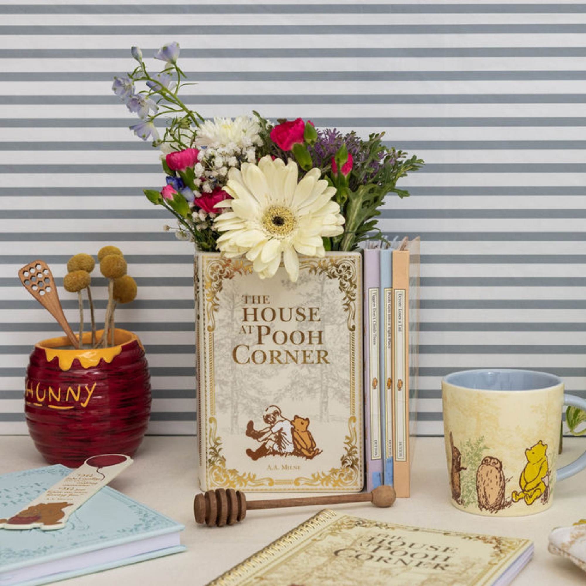 Decorative setup with 'The House at Pooh Corner' book, flowers, and Winnie the Pooh-themed items against a striped background.