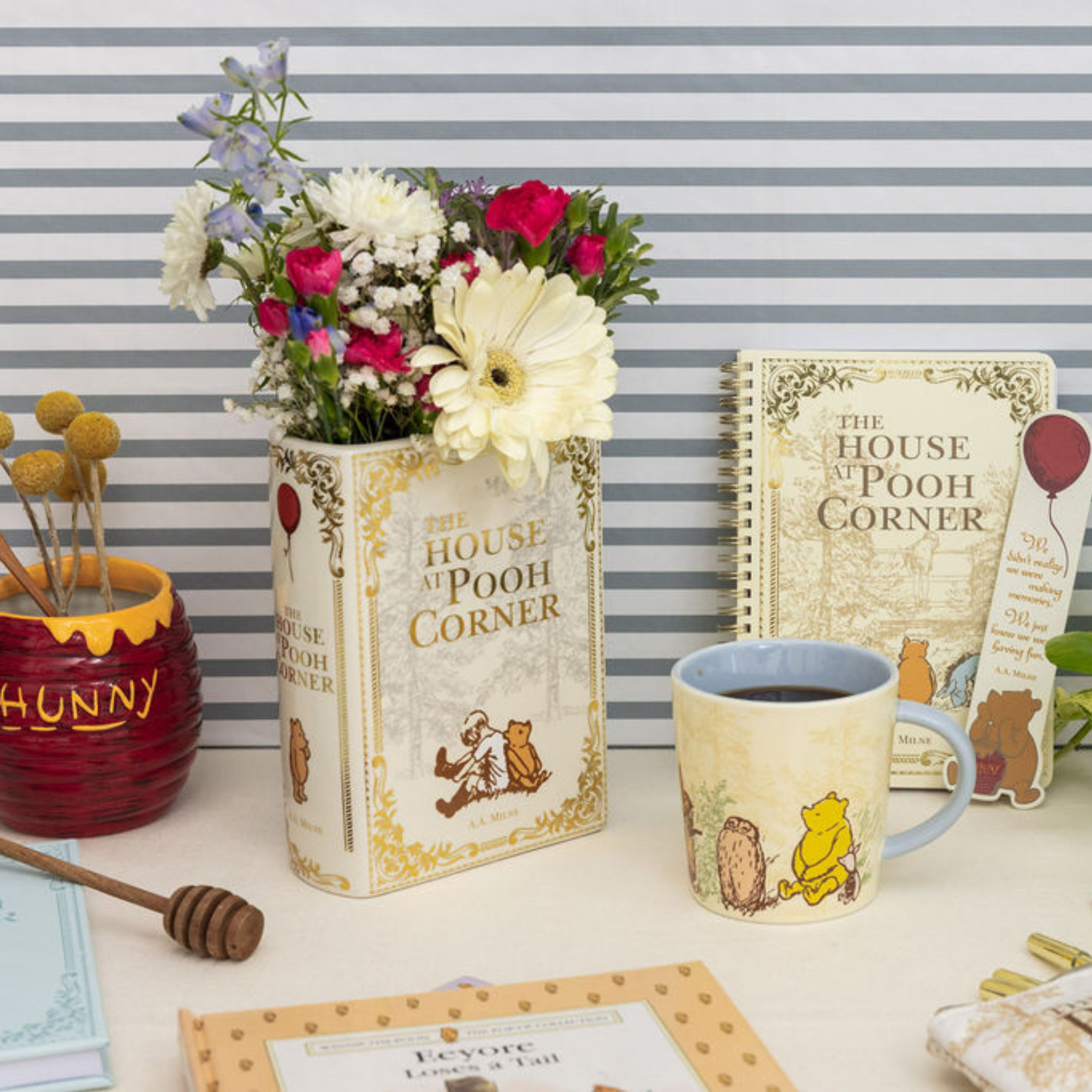 Decorative setup with 'The House at Pooh Corner' book, mug, flowers, and honey pot on a striped background.