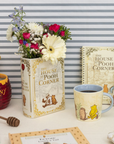 Decorative setup with 'The House at Pooh Corner' book, mug, flowers, and honey pot on a striped background.