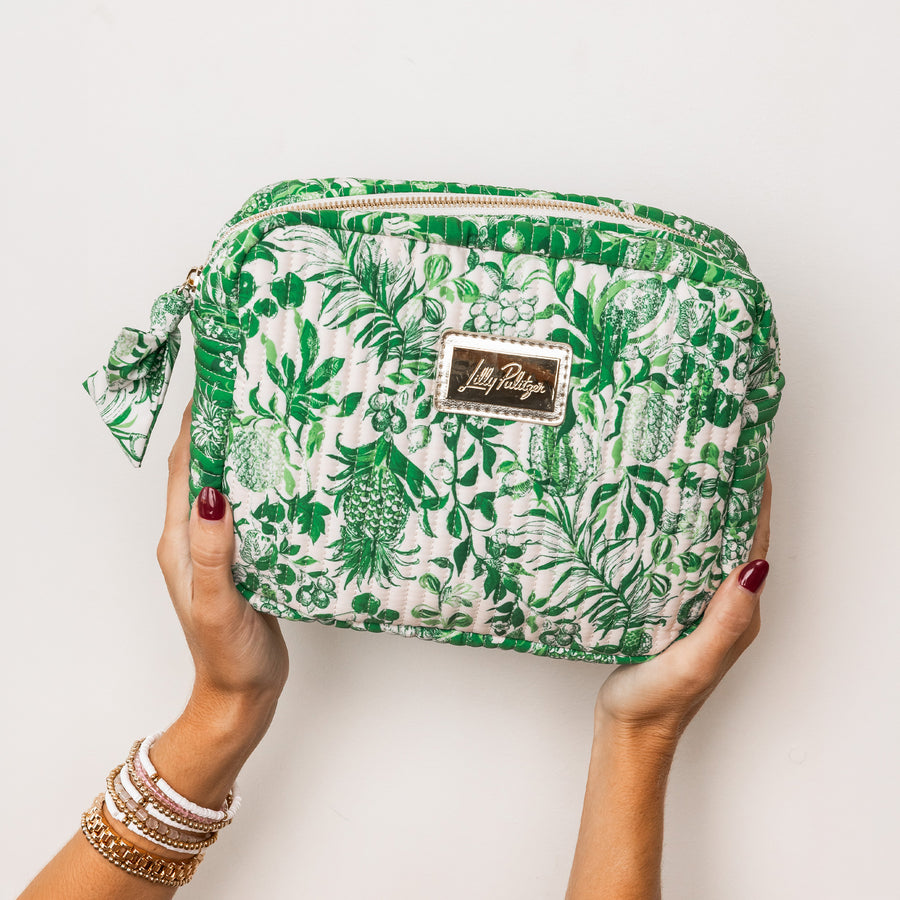 model holding rectangular zippered pouch with a green tropical botanical print on a cream background, featuring a small gold-tone metal brand plaque on the front and a matching fabric wristlet strap on the left side.