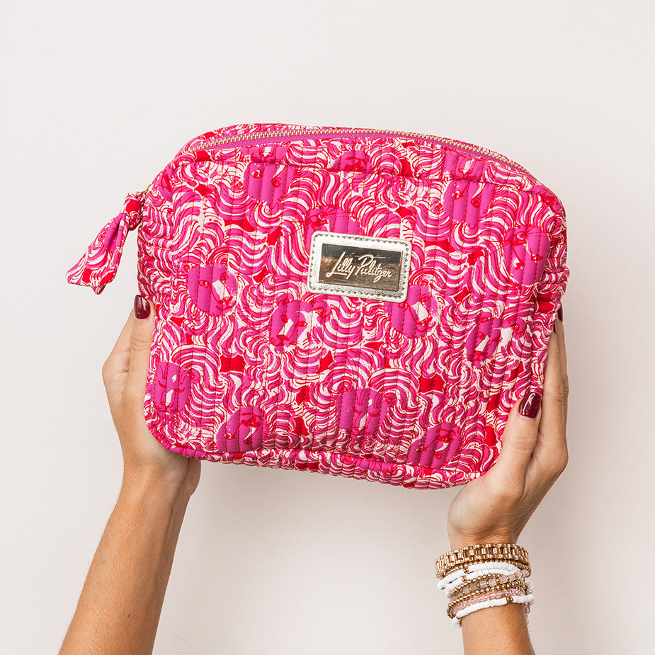 model holding bright pink cosmetic pouch with a white and red abstract pattern, featuring a gold-tone metal brand plaque on the front and a matching zipper pull.