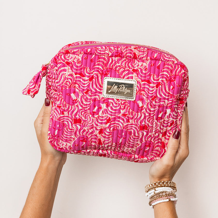 model holding bright pink cosmetic pouch with a white and red abstract pattern, featuring a gold-tone metal brand plaque on the front and a matching zipper pull.