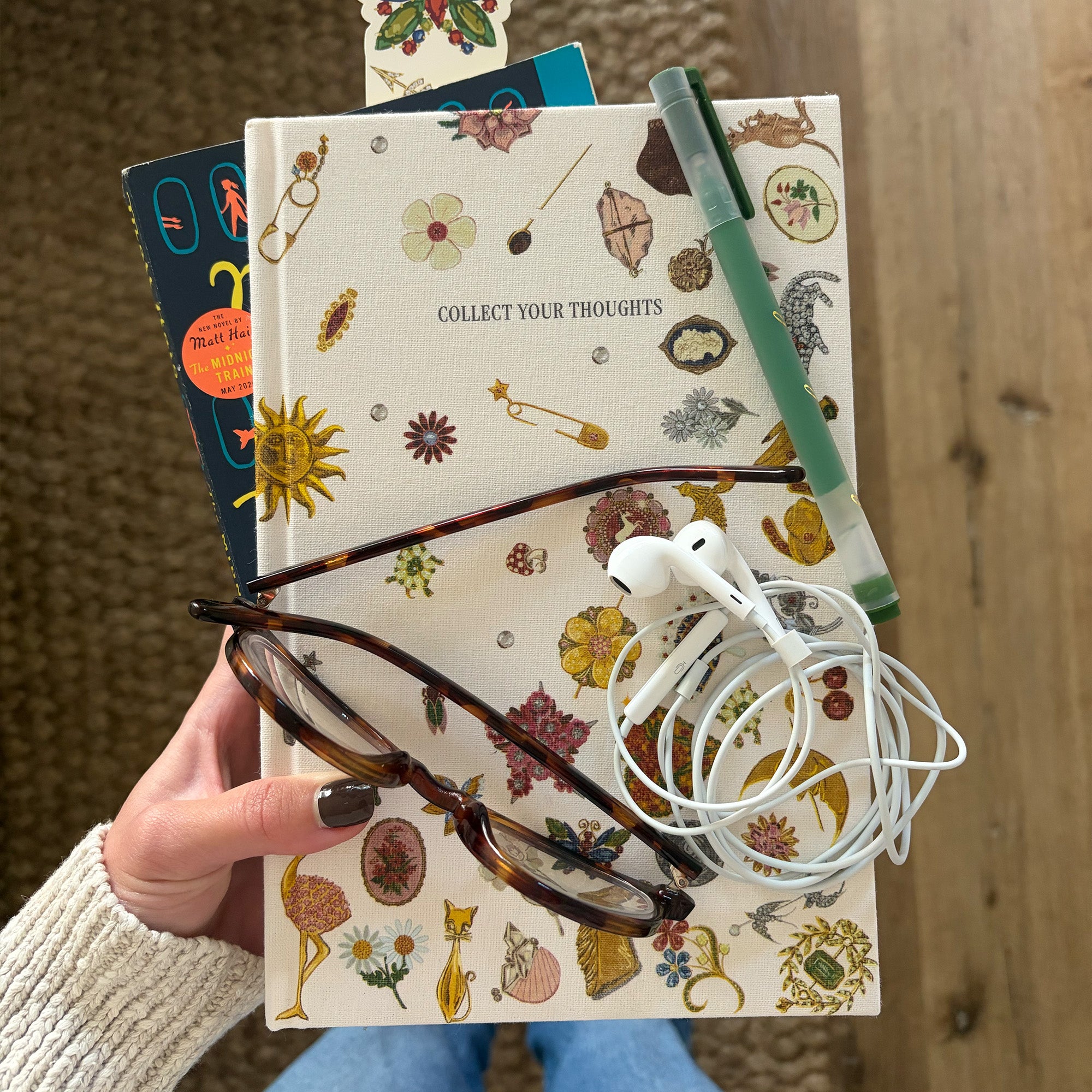 model holding a notebook with a cream-colored cover decorated with various whimsical illustrations such as a sun, flamingo, flowers, and tools, featuring the text 'COLLECT YOUR THOUGHTS' and a blue ribbon bookmark.