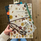 model holding a notebook with a cream-colored cover decorated with various whimsical illustrations such as a sun, flamingo, flowers, and tools, featuring the text 'COLLECT YOUR THOUGHTS' and a blue ribbon bookmark.