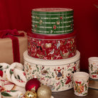 Three stacked round Christmas-themed metal tins with matching holiday cups, alongside a wrapped gift box and ornaments, set against a dark red background.