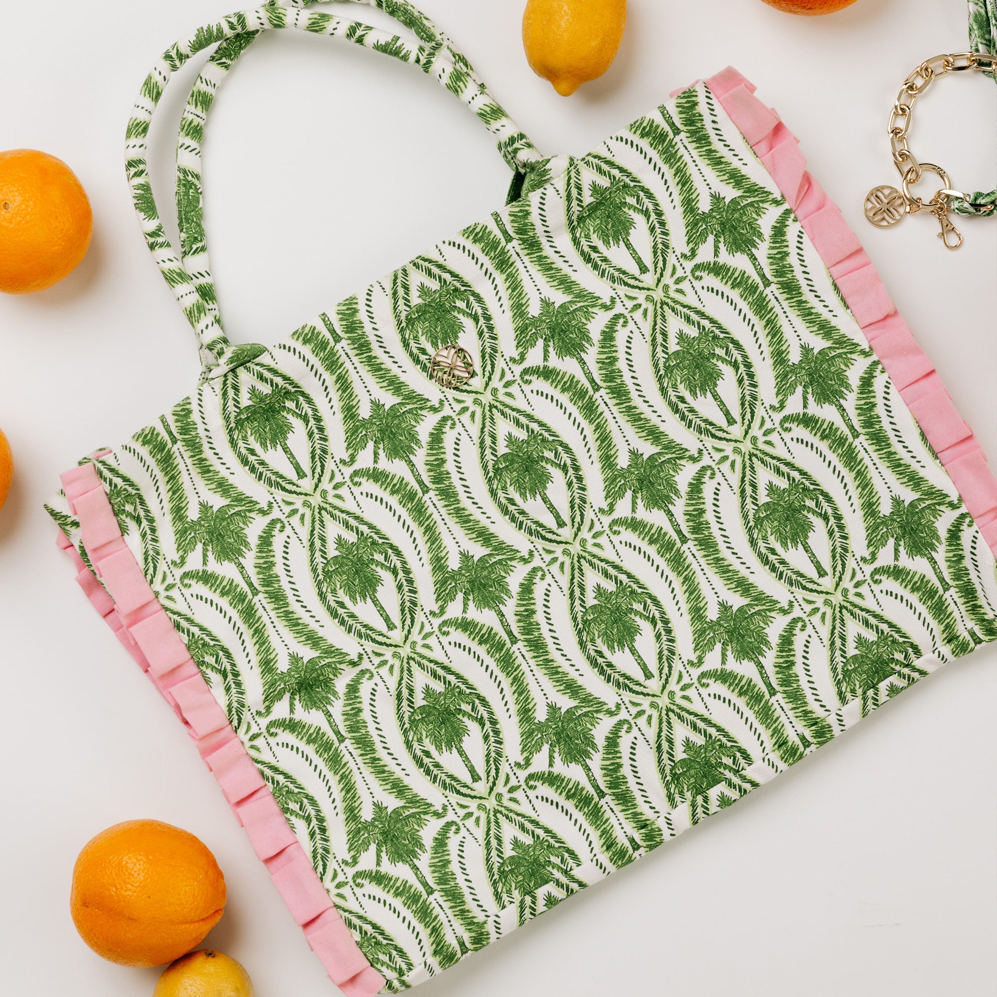 A tote bag with a green palm tree pattern on a white background, finished with pink ruffle trim along the edges and handles. The bag is styled in a flat-lay composition with lemons and oranges on a white background.