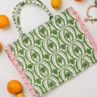 A tote bag with a green palm tree pattern on a white background, finished with pink ruffle trim along the edges and handles. The bag is styled in a flat-lay composition with lemons and oranges on a white background.