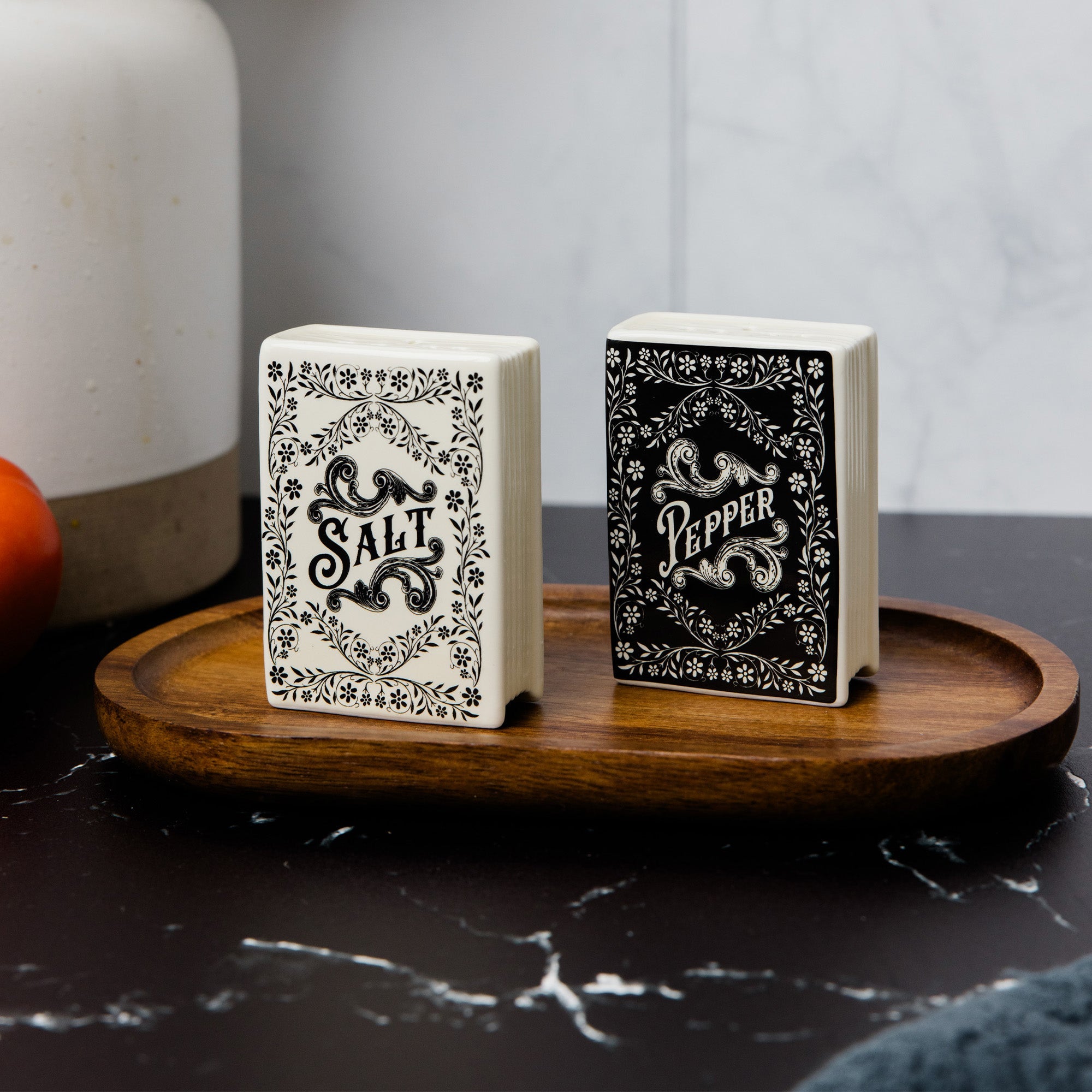 Two decorative playing card boxes labeled SALT and PEPPER, standing upright on a round wooden tray. The salt box is off-white with black ornate lettering and border, and the pepper box is black with white ornate lettering and border.