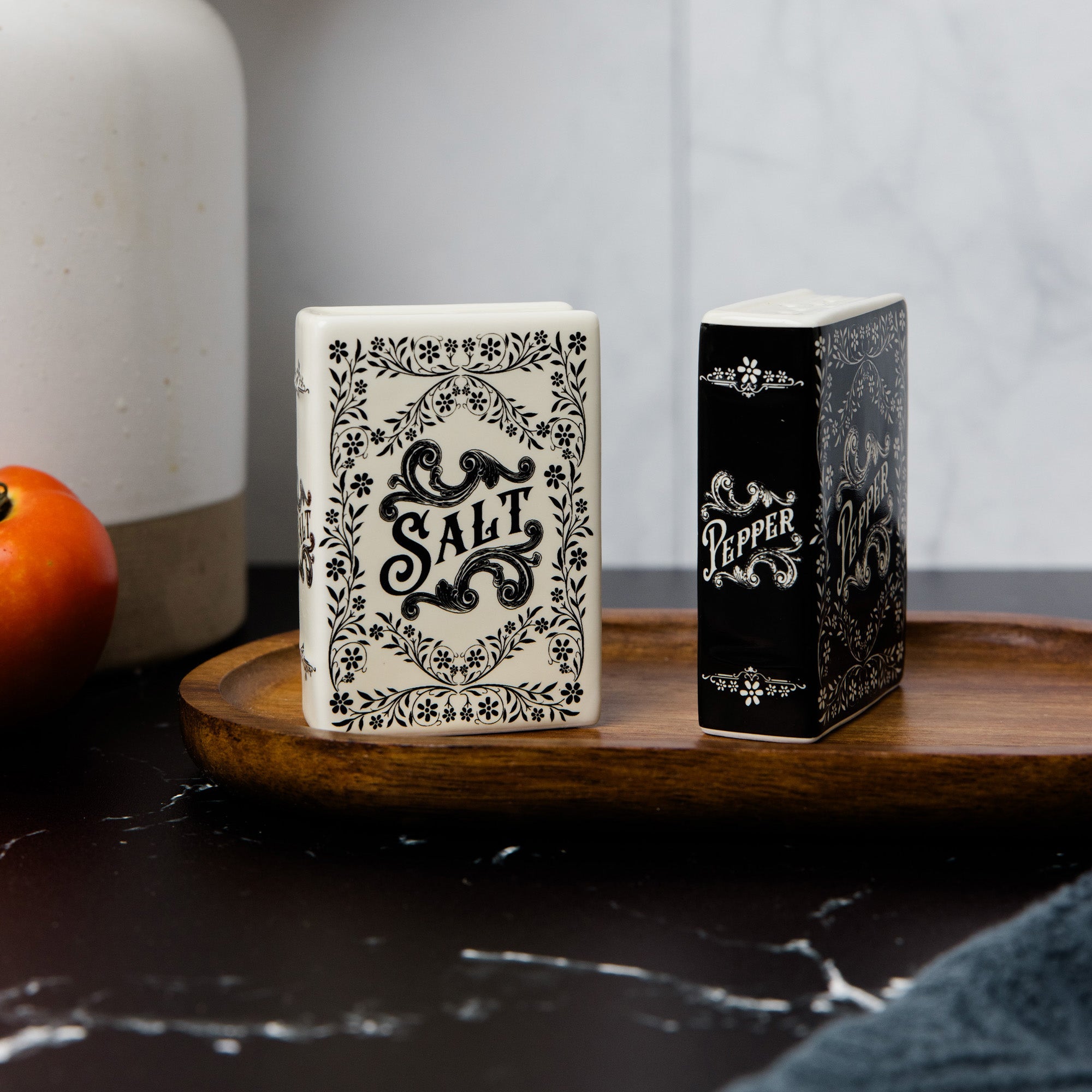 A decorative ceramic salt and pepper shaker set on a wooden tray. The salt shaker is off-white with black floral scrollwork and the word 'SALT', while the pepper shaker is black with silver-gray floral scrollwork and the word 'PEPPER'.