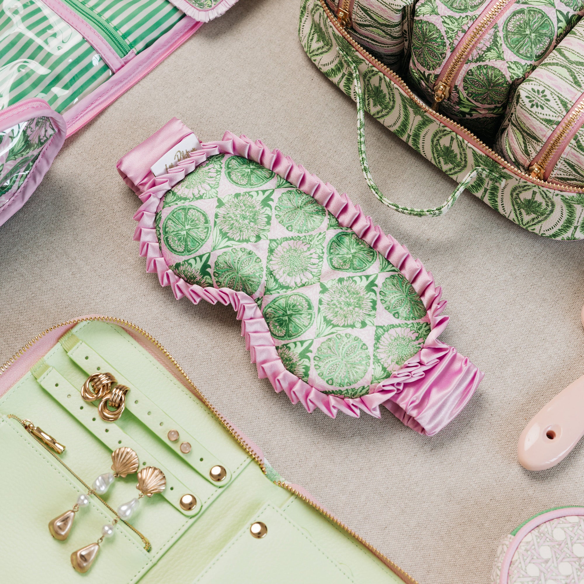 A styled flat-lay of a pink and green patterned sleep mask in the center, surrounded by matching green and pink patterned accessories on a neutral beige surface.