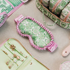 A styled flat-lay of a pink and green patterned sleep mask in the center, surrounded by matching green and pink patterned accessories on a neutral beige surface.