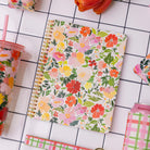 Floral notebook on a tiled surface with a pink tumbler and other stationery items.