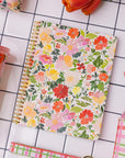 Floral notebook on a tiled surface with a pink tumbler and other stationery items.