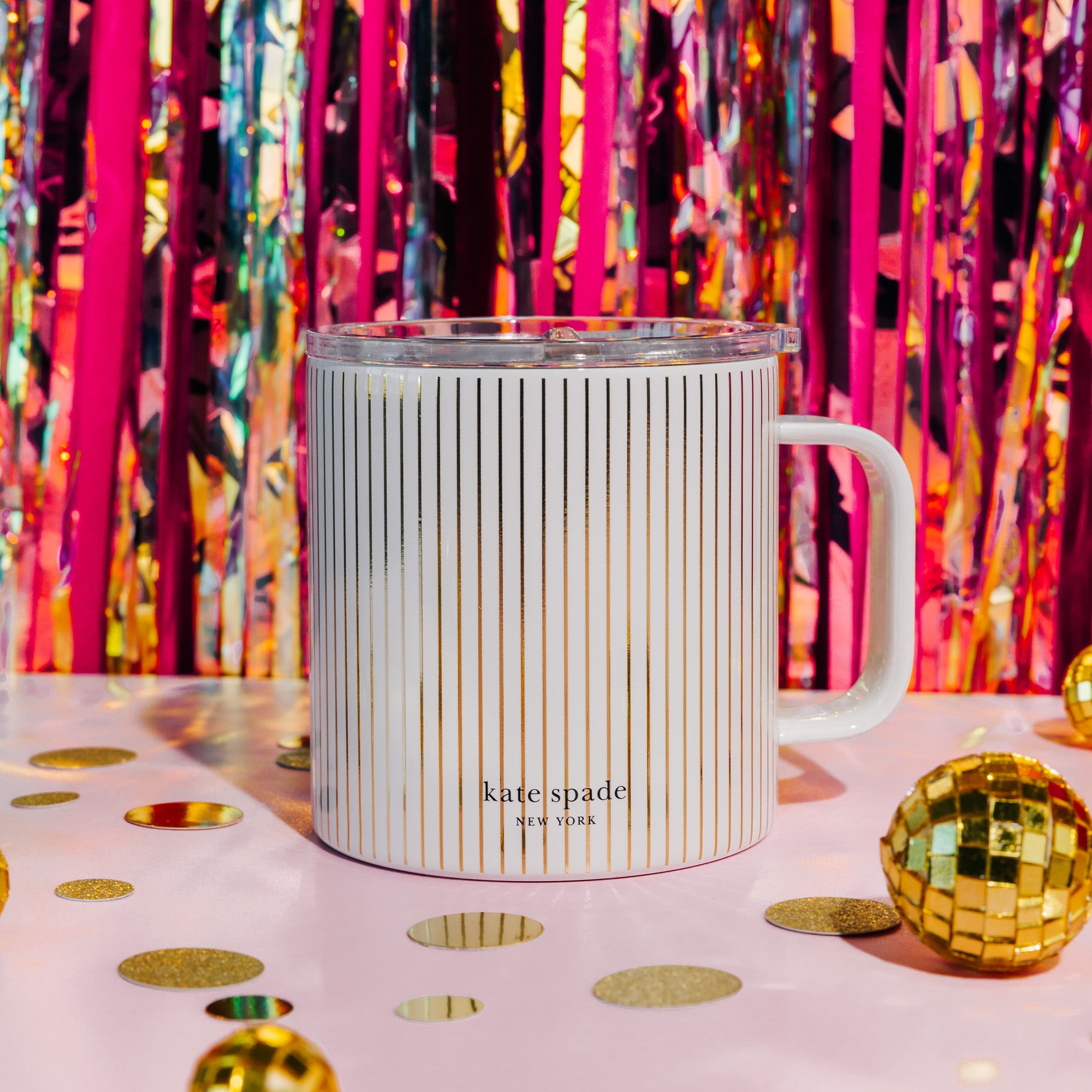 A Kate Spade New York mug with a white handle, featuring a cream and gold vertical stripe pattern, a clear plastic lid, and the brand name printed near the bottom.