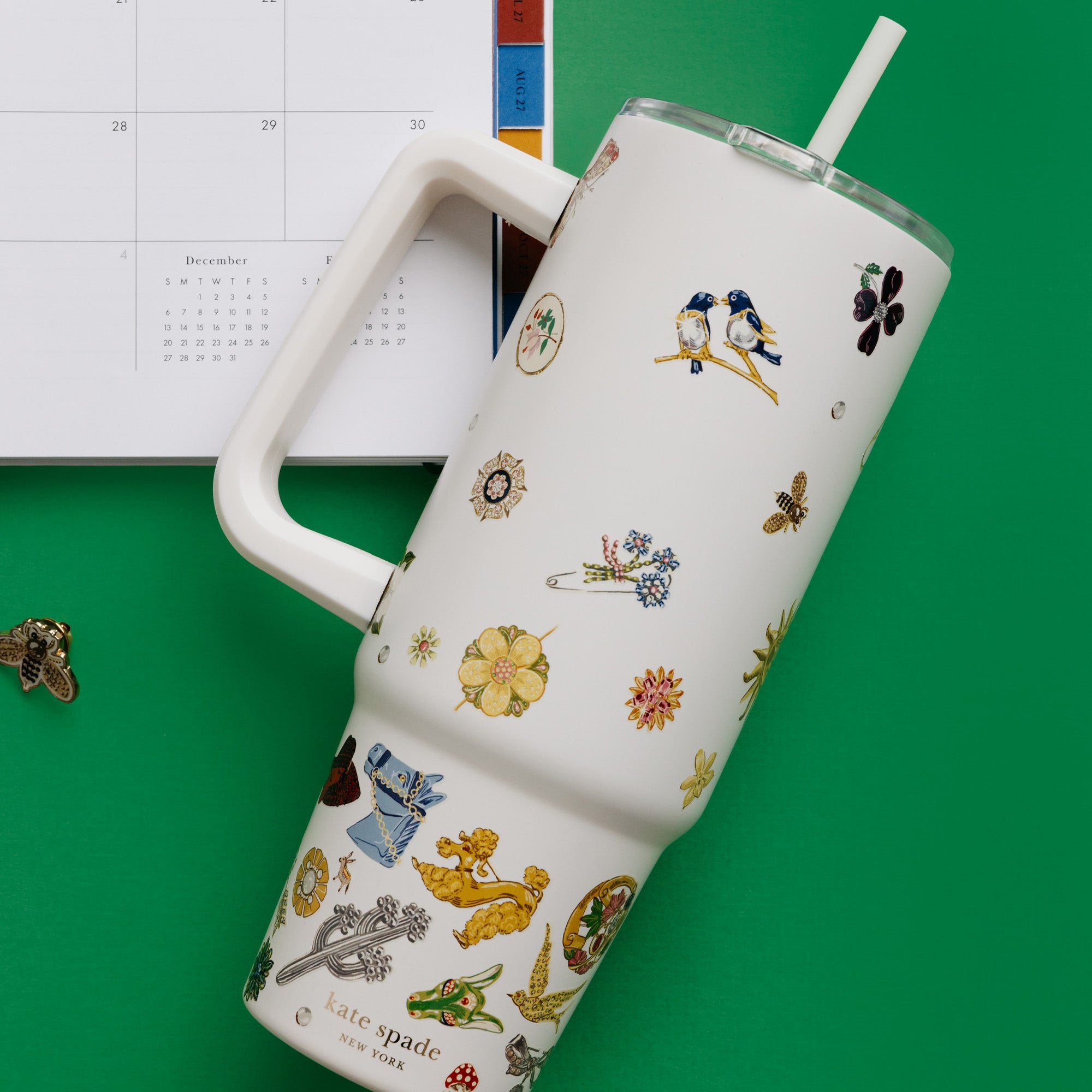 An insulated travel tumbler with a handle, clear lid, and straw, decorated with a whimsical pattern of small birds, flowers, and nature illustrations on a warm cream background.
