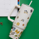 An insulated travel tumbler with a handle, clear lid, and straw, decorated with a whimsical pattern of small birds, flowers, and nature illustrations on a warm cream background.