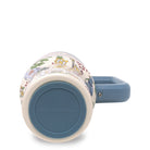 tumbler that features a cream-colored body with a detailed multicolor illustration of a garden scene, a solid blue handle, and a matching blue circular base insert with the brand name debossed.