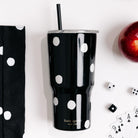 A black stainless steel tumbler with white polka dots and a black straw, featuring the gold 'kate spade NEW YORK' logo near the base. The tumbler is styled in a flat-lay arrangement on a white background alongside a matching polka-dot cloth, dice, playing cards, and a red apple.