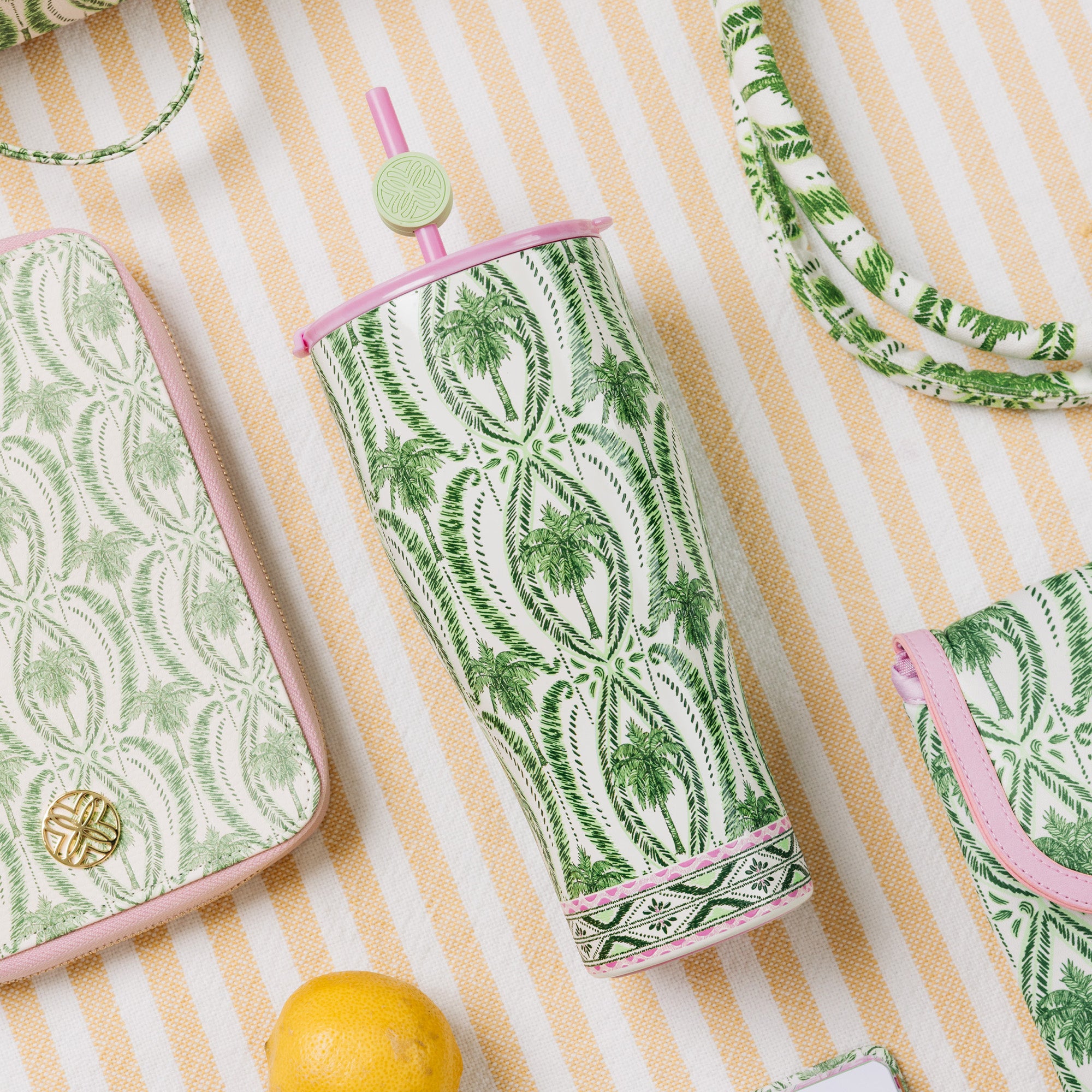 A green and white palm tree patterned insulated tumbler with a pink straw and pink rim, styled on a yellow and white striped fabric alongside matching accessories.