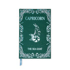 A dark green hardcover notebook or journal featuring a silver-toned Capricorn mermaid illustration and decorative border, with a light blue ribbon bookmark hanging from the bottom.
