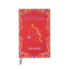 A red fabric-covered notebook or journal featuring the Sagittarius zodiac design, with a purple ribbon bookmark hanging from the bottom edge. The cover has a decorative border and shows the text 'SAGITTARIUS' and 'THE ARCHER'.