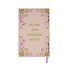 A pink notebook with a decorative border and gold text reading 'ANALYTICAL CLEVER COMPASSIONATE MAGNETIC' on the cover, with a purple ribbon bookmark hanging from the bottom right.
