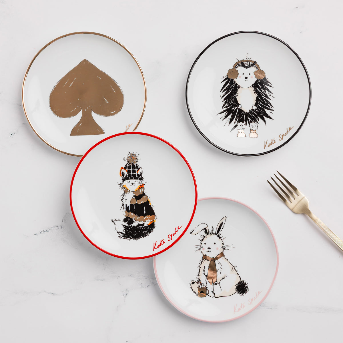 Scattered four round white plates on a white background, each with a different illustrated character and border color: a white rabbit with a pink rim, a brown fox with a red rim, a hedgehog with a black rim, and a large gold spade symbol with a gold rim. The name 'Kate Spade' appears in script near each plate.