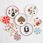 Scattered four round white plates on a white background, each with a different illustrated character and border color: a white rabbit with a pink rim, a brown fox with a red rim, a hedgehog with a black rim, and a large gold spade symbol with a gold rim. The name 'Kate Spade' appears in script near each plate surrounded by cocktail napkins, candy and coasters