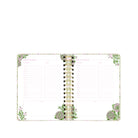 An open spiral-bound planner with decorative floral corner illustrations and a gold-and-black spiral binding, shown on a plain white background.