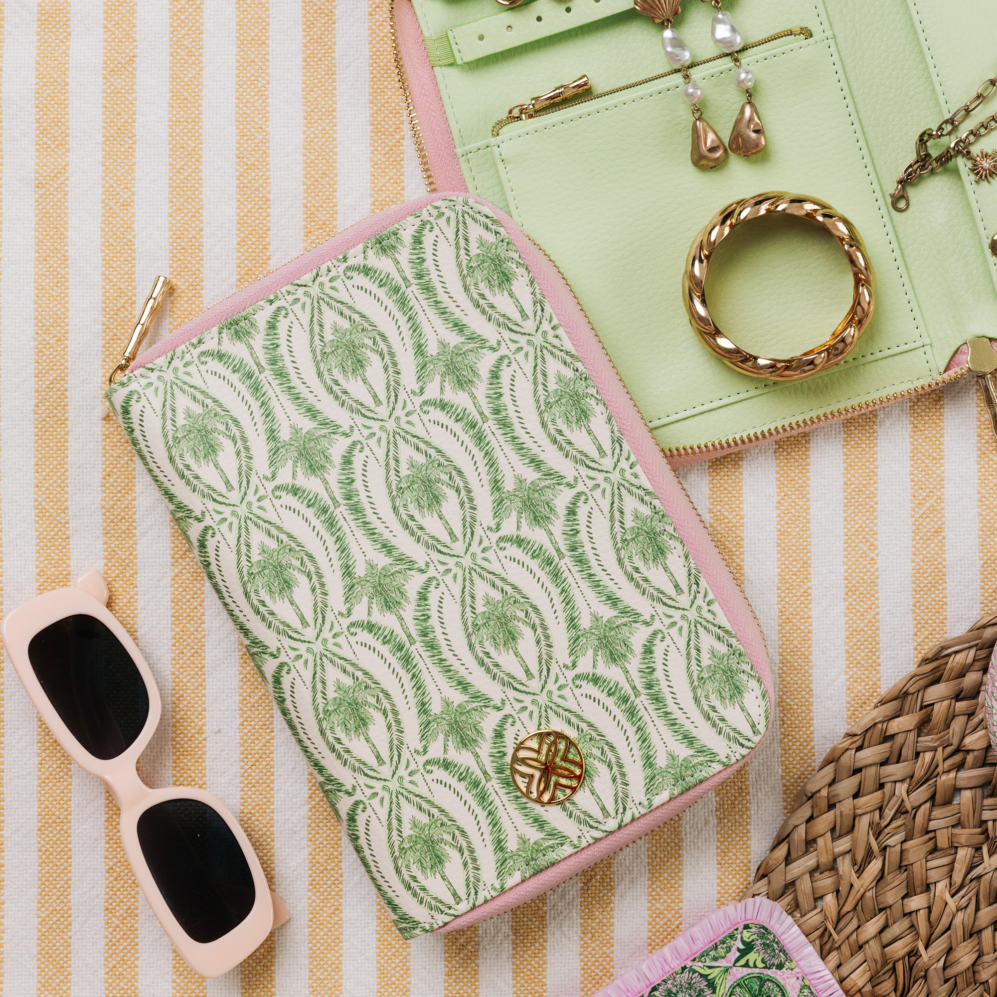 A flat-lay product photo of a zippered pouch with a green palm tree pattern on a cream background and light pink trim, styled with sunglasses, a mint green jewelry organizer, and a woven bag on a yellow and white striped blanket.