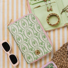 A flat-lay product photo of a zippered pouch with a green palm tree pattern on a cream background and light pink trim, styled with sunglasses, a mint green jewelry organizer, and a woven bag on a yellow and white striped blanket.
