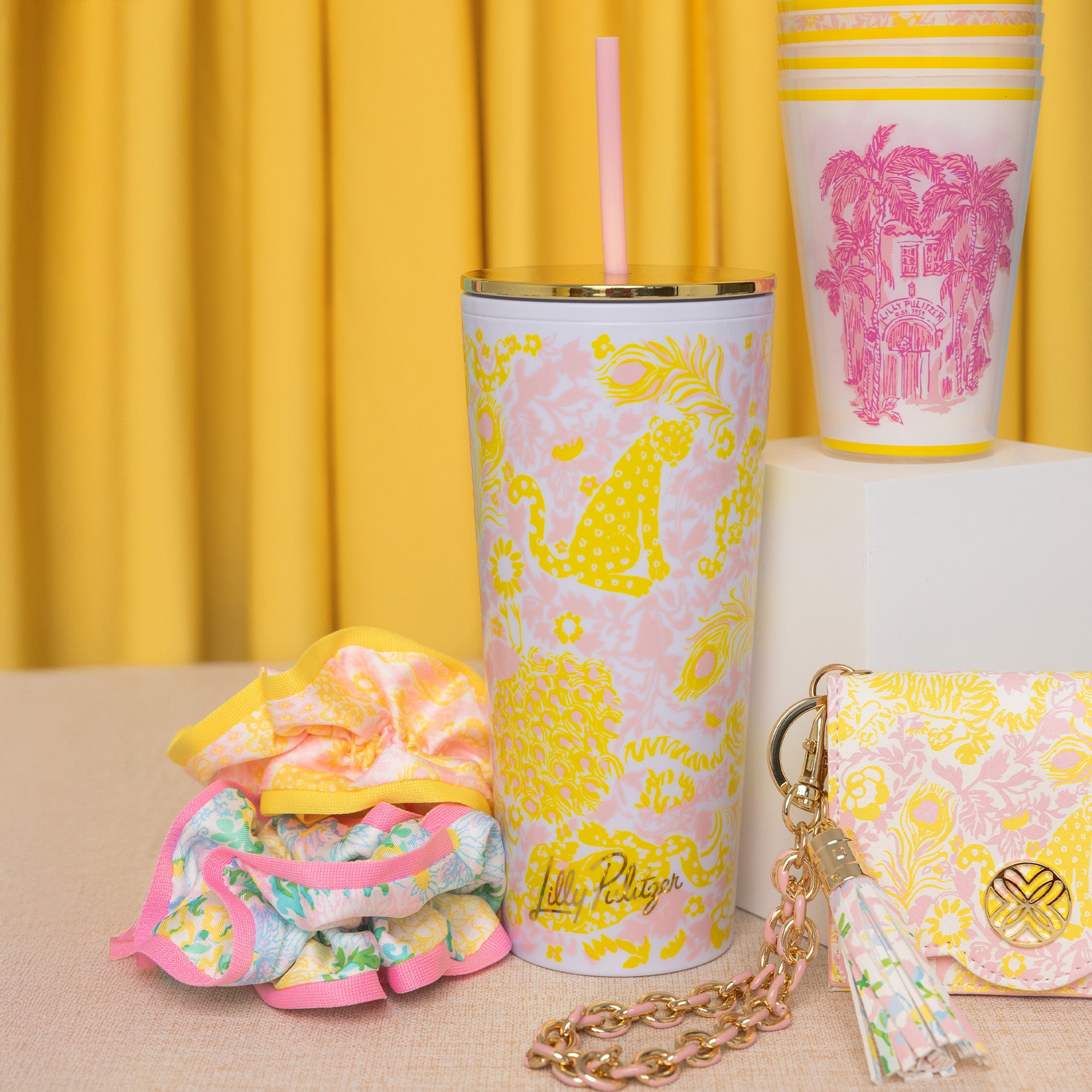 Colorful tumbler, keychain, and pouch with pink and yellow patterns on a yellow curtain background.