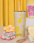 Colorful tumbler, keychain, and pouch with pink and yellow patterns on a yellow curtain background.
