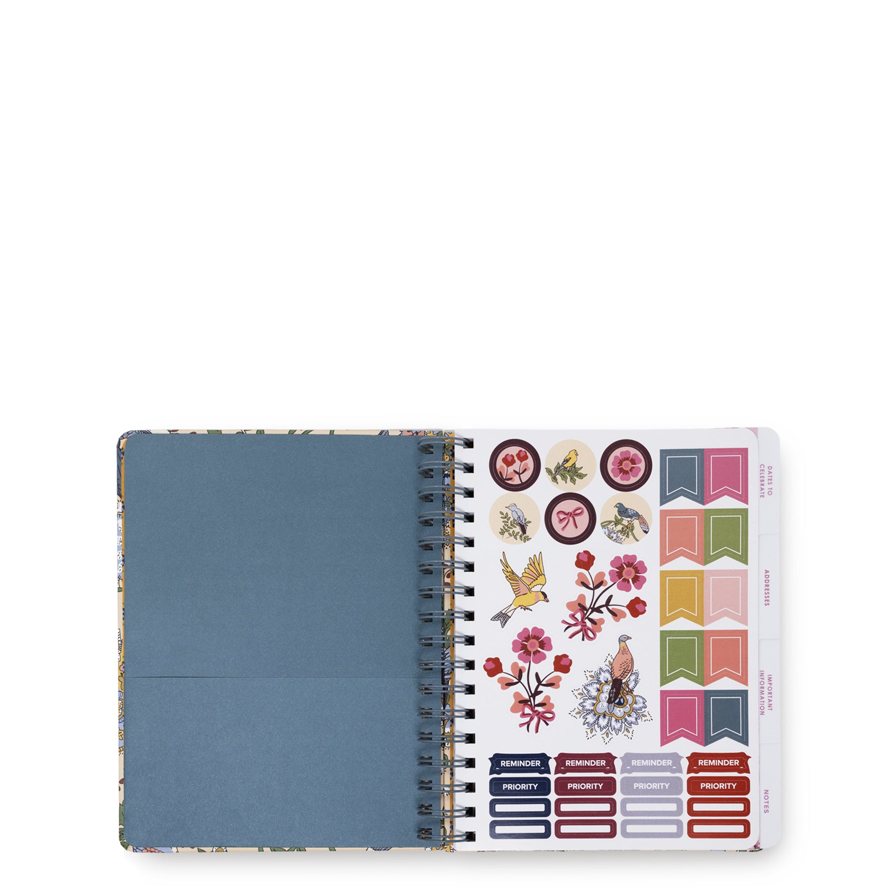An open spiral-bound planner or notebook with a dusty blue left page and a sticker sheet on the right featuring colorful decorative stickers, flags, labels, and floral designs, photographed on a white background.