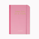 A pink hardcover planner with a matching pink elastic closure band. The front cover features the text 'ONE DAY AT A TIME' in a gold metallic font, with the brand name 'as it's planned to be' printed in small white script near the bottom.