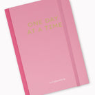 A pink hardcover planner with gold foil text reading 'ONE DAY AT A TIME' and a small phrase 'as it's planned to be' near the bottom, shown on a clean white background.