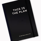 A black journal with white text reading 'THIS IS THE PLAN' and a smaller phrase near the bottom, shown on a clean white background.