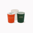 Three cylindrical candle tins with matching lids are arranged on a white background. The left tin is orange with a white lid featuring a winterberry illustration and the text 'A 1925 WINTERBERRY.