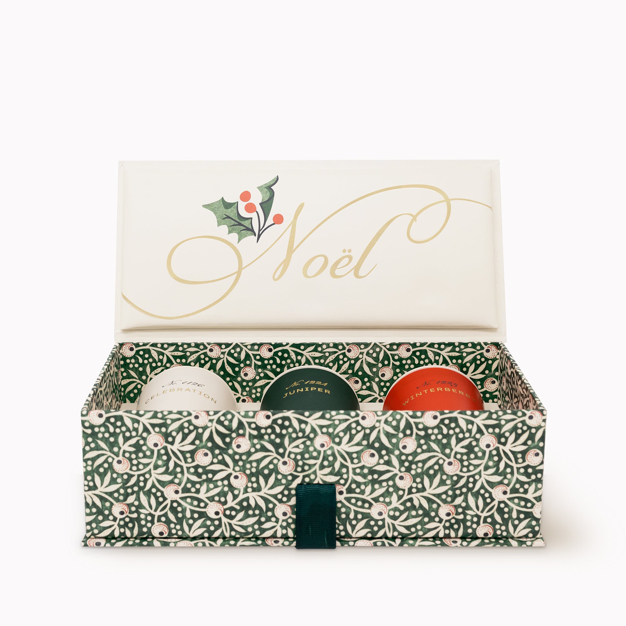 An open Christmas gift box with a cream interior and lid decorated with gold 'Noël' script and a holly illustration. The box has a green and cream floral pattern, and three round candles are partially visible inside.