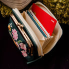 A dark teal floral zip-around wallet with a gold-tone kiss-lock clasp, shown open with several cards inserted. The wallet is photographed on a dark surface with decorative props in the background.