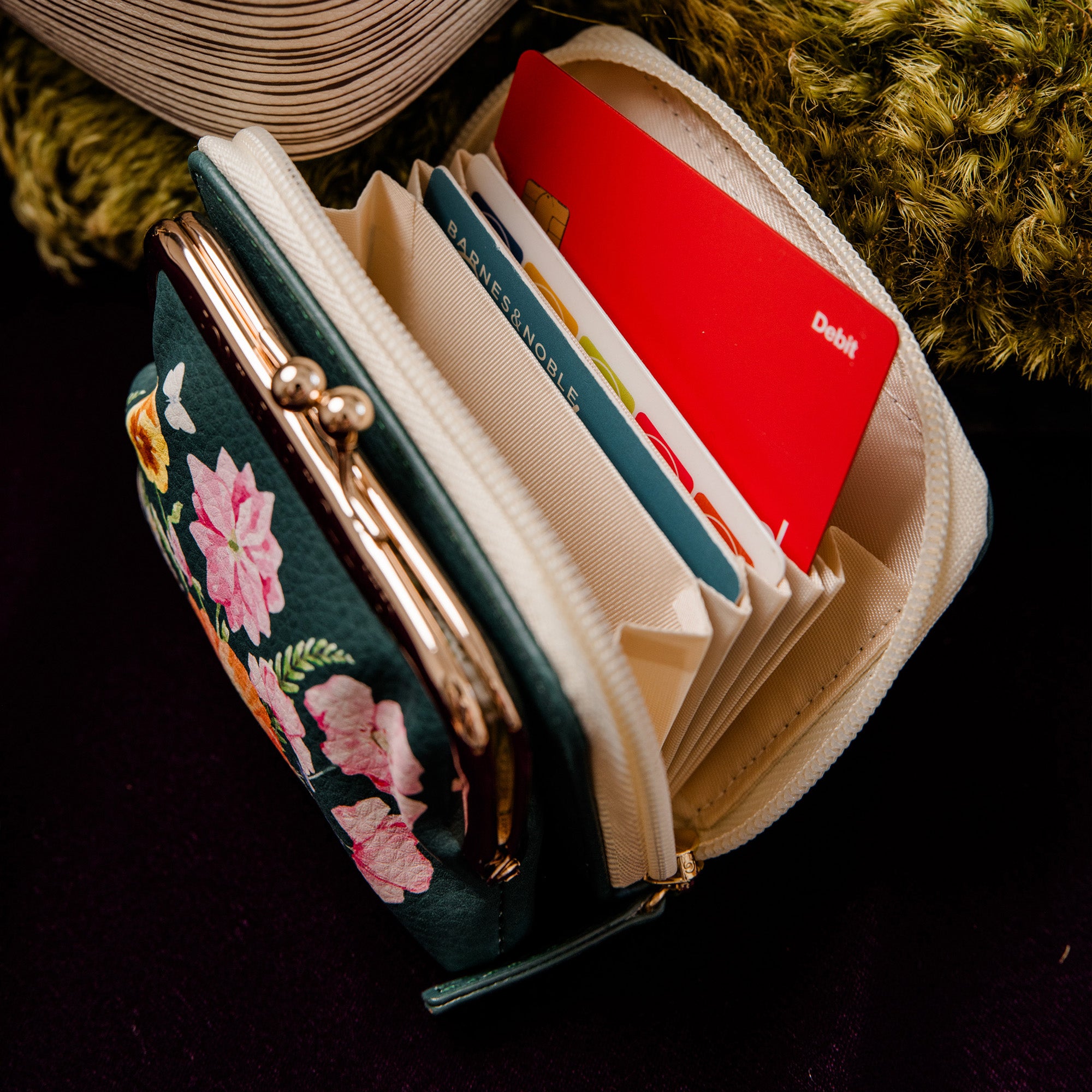 A dark teal floral zip-around wallet with a gold-tone kiss-lock clasp, shown open with several cards inserted. The wallet is photographed on a dark surface with decorative props in the background.