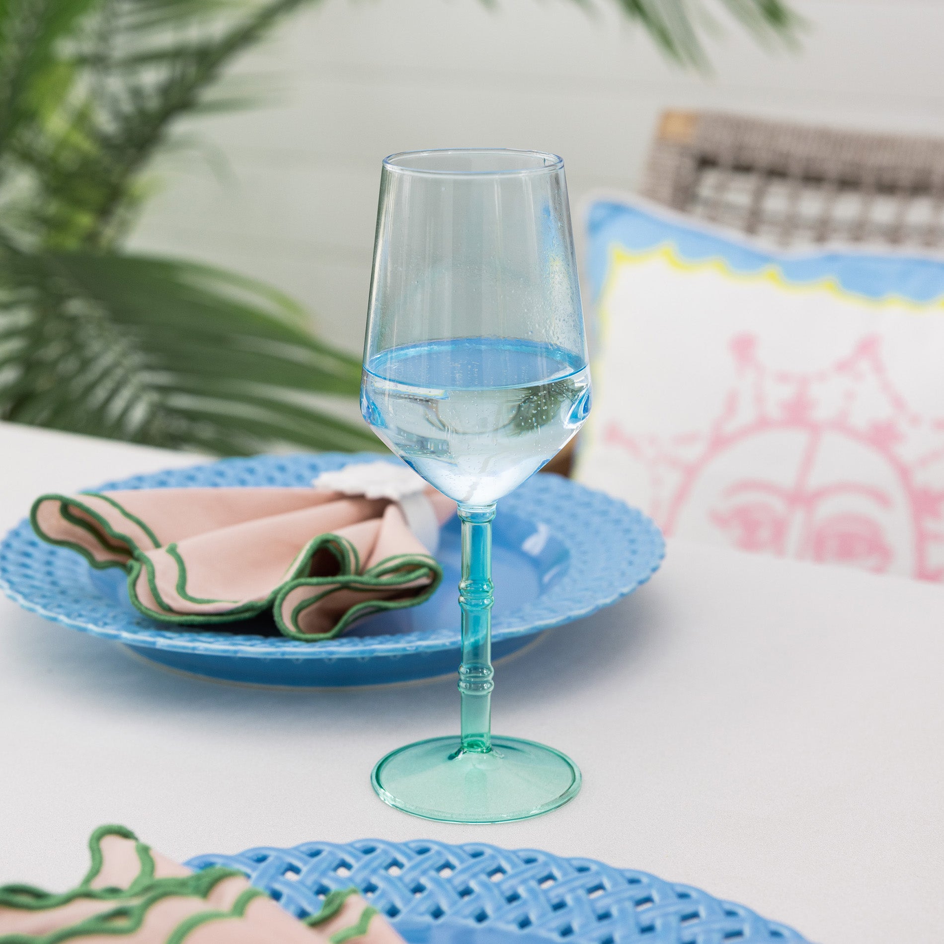 Clear wine glass with blue water on a blue plate with a colorful napkin, set against a blurred outdoor background.