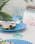 Clear wine glass with blue water on a blue plate with a colorful napkin, set against a blurred outdoor background.