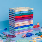 A stack of colorful zodiac-themed notebooks or journals with pastel covers and printed zodiac names, arranged on a light blue surface with matching zodiac-shaped accessories scattered around.