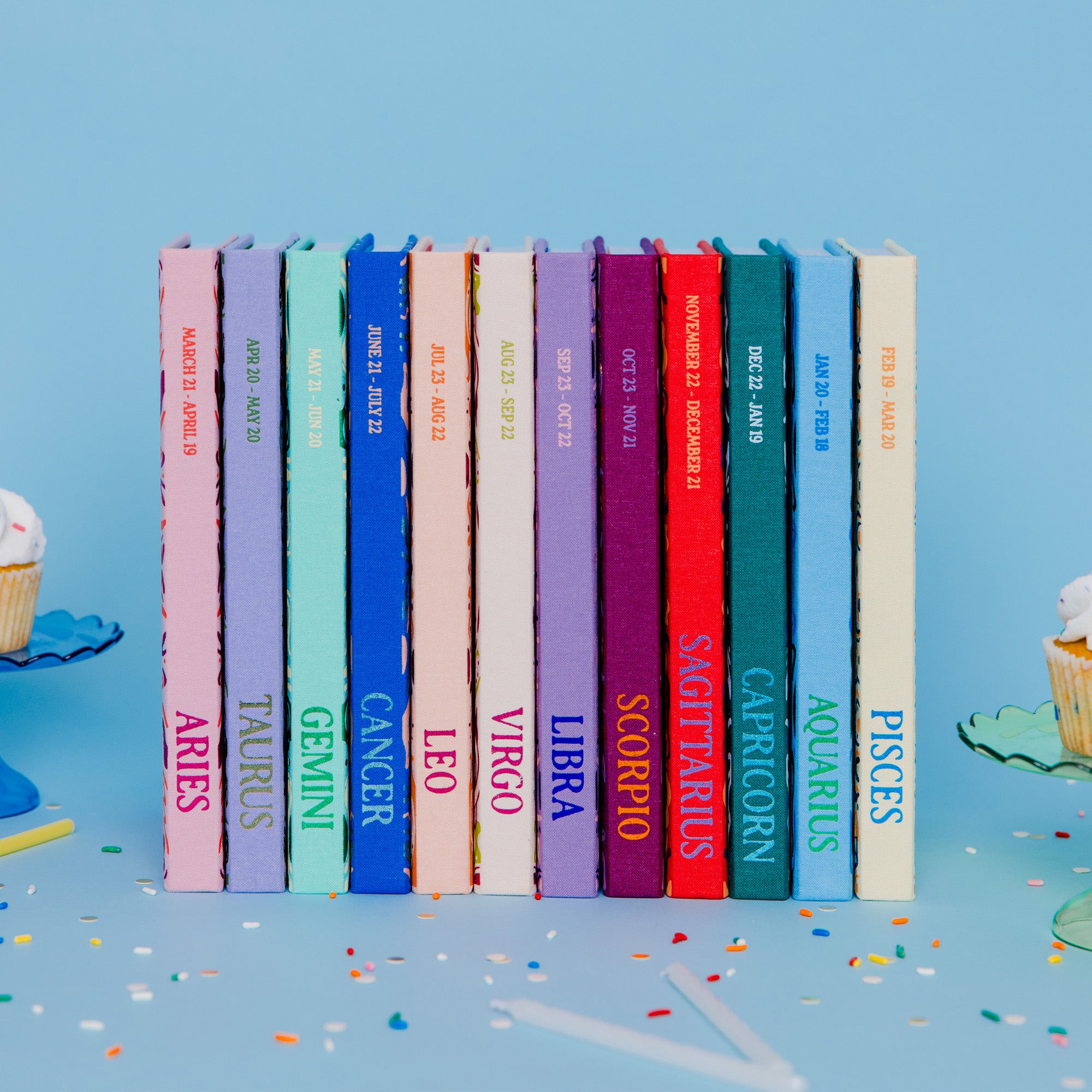 A set of twelve hardcover books arranged in a row, each with a different colored spine representing a zodiac sign, set against a light blue background with decorative cupcakes on the sides.