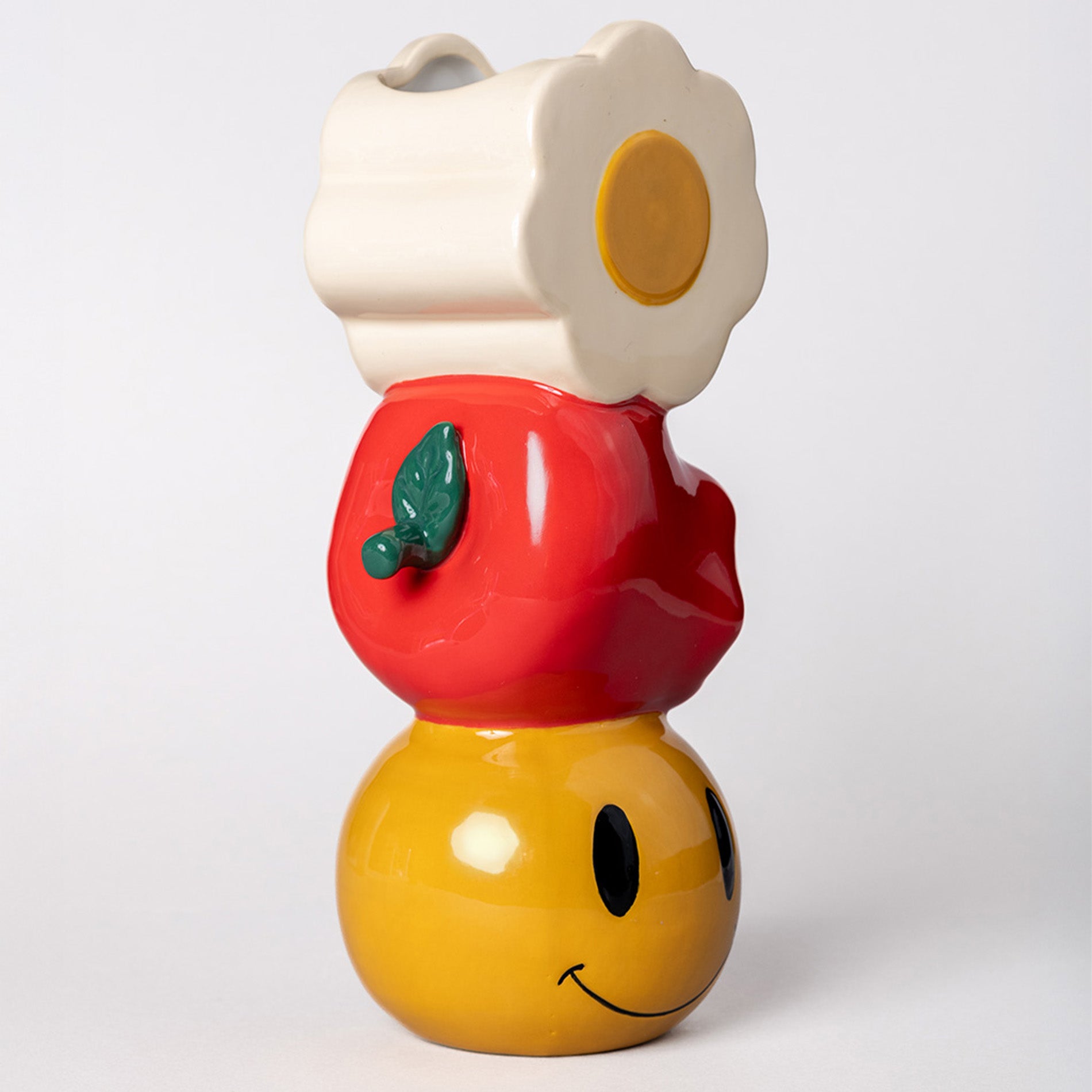 Colorful ceramic vase with stacked white daisy, apple, and yellow smiley on a white background.